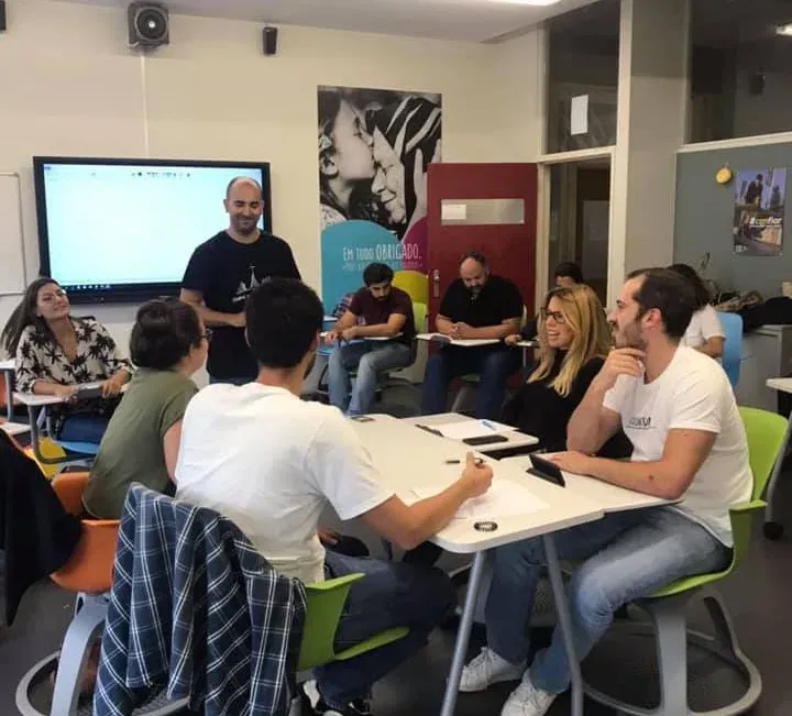 A facilitator stands by a screen while participants seated around classroom tables listen, smiling